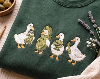 May include: A dark green crewneck sweatshirt featuring an embroidered design of four white ducks interacting with pickles. One duck holds a jar of pickles, one wears a pickle costume, one holds a stack of pickle slices, and one holds a single pickle.