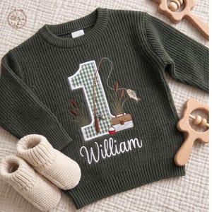 May include: A dark olive green long-sleeve knit sweater featuring an embroidered number one with a green and white gingham pattern. The design includes a fishing rod, a tackle box, and the name William in white cursive script lettering.
