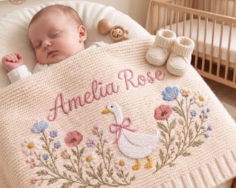 Puede incluir: Una manta de punto color crema para bebé con el nombre Amelia Rose bordado en rosa, una oca blanca central con un lazo rosa y bordados florales en tonos rosa, azul y blanco, acompañada de patucos de bebé en color crema.