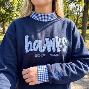 May include: A navy blue sweatshirt featuring a mock neck collar with a blue and white gingham pattern. The word hawks is appliqued in blue and white gingham across the chest, with SCHOOL NAME embroidered below and Jennifer with a heart on the sleeve.