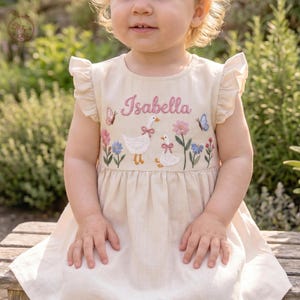 May include: A beige sleeveless dress with ruffled shoulders featuring embroidered details of two white geese with pink bows, colorful flowers, and butterflies. The name Isabella is embroidered in pink script text across the chest area.
