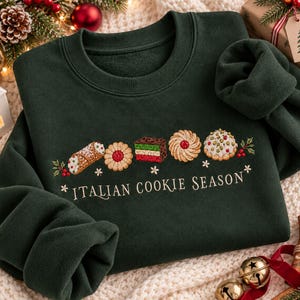 May include: A dark forest green crewneck sweatshirt featuring an embroidered row of various Italian cookies and the text "ITALIAN COOKIE SEASON" in a cream-colored font. The design includes a cannoli, a jam cookie, a layered cake, and others.