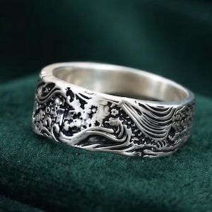 Puede incluir: Un anillo de banda de plata con un intrincado diseño en relieve de olas oceánicas arremolinadas y pequeños elementos florales. El metal tiene un acabado pulido en las superficies elevadas y un acabado oscuro y oxidado en las zonas hundidas.