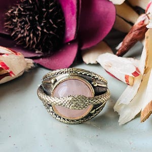 May include: A close-up of a ring featuring a pale pink gemstone at its centre. The ring is adorned with a textured, gold-toned snake motif. The background includes a deep purple flower and dried, cream-coloured petals.
