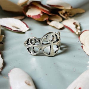 Puede incluir: Un anillo plateado con un diseño calado. El anillo presenta dos formas circulares conectadas, cada una con un patrón en forma de cruz en el interior. El anillo está sobre una superficie azul claro con pétalos secos dispersos.