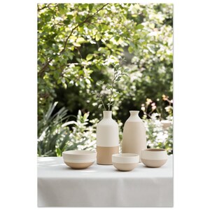 Può includere: Una collezione di vasi e ciotole in ceramica in una palette di colori neutri. I vasi sono alti e cilindrici, con un design bicolore. Le ciotole sono rotonde e hanno una combinazione di colori simile. Gli oggetti sono disposti su una tovaglia bianca con uno sfondo verde sfocato.