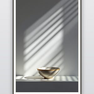 May include: A framed wall art print featuring a light grey bowl with a gold rim, centred on a white surface. The composition includes dramatic diagonal shadows cast across the wall and surface, with the text MULTIPLE SIZES · INCHES & CM below.