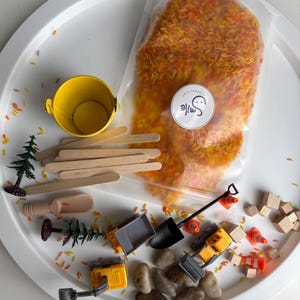 Peut inclure: Un kit de jeu sensoriel comprenant un sachet transparent de riz teint en orange et jaune avec un autocollant Smile Sensory Play, un seau en métal jaune, des bâtonnets en bois, une pelle en bois, deux engins de chantier en plastique, des cailloux.