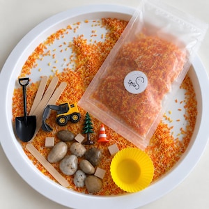 Peut inclure: Un plateau circulaire blanc contenant du riz teint en orange et jaune, un sachet en plastique transparent avec le logo SmieS, une pelle en plastique noir, des bâtonnets en bois, une pelleteuse jouet jaune, des galets gris, des cubes en bois, un sapin, un cône et un gobelet jaune.
