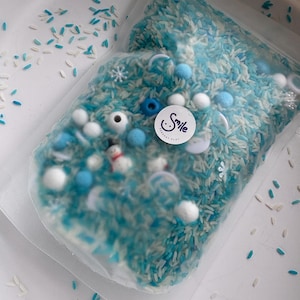 Peut inclure: Un sachet en plastique transparent rempli d'un mélange de riz teint en blanc et bleu, de petits pompons blancs et bleus, de formes de flocons de neige blancs, de perles bleues et d'une petite figurine de bonhomme de neige. Un autocollant Smile Sensory Play.