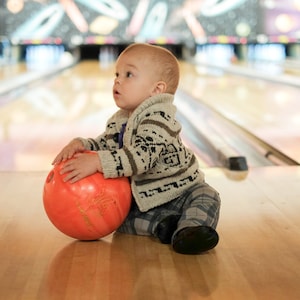 Può includere: Un bambino seduto su una pista da bowling tiene in mano una palla da bowling arancione. Il bambino indossa un maglione fantasia, pantaloni a quadri e scarpe nere. Le piste e i birilli da bowling sono visibili sullo sfondo.