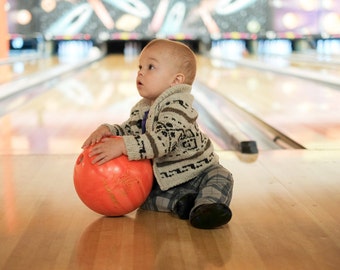 May include: A baby sits on a bowling lane, holding an orange bowling ball. The baby wears a patterned sweater, plaid pants, and black shoes. The bowling alley lanes and pins are visible in the background.