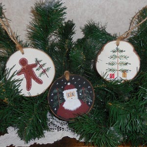 May include: Three rustic Christmas ornaments. One features a gingerbread man, another a Christmas tree, and the third Santa Claus. Each ornament is a wooden slice with a twine loop for hanging. The ornaments are displayed on a bed of evergreen branches.