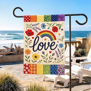 Puede incluir: Una bandera de jardín rectangular con un centro color crema que presenta la palabra love en letra cursiva azul oscuro, un arcoíris y varias ilustraciones florales coloridas. Los bordes superior e inferior tienen seis parches de tela.