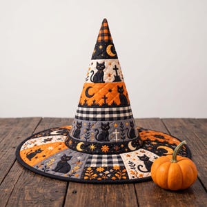 May include: A quilted witch hat featuring a pattern of black cats, crescent moons, stars, and crosses in orange, black, and white. The conical hat has a wide brim and a checkered top section, accompanied by a small orange pumpkin placed nearby.