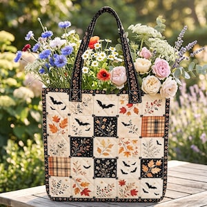 May include: A quilted tote bag featuring a patchwork design of cream and black squares decorated with autumn leaves, branches, and flying bats. The bag has two black fabric handles and is filled with a colorful bouquet of fresh garden flowers.