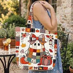May include: A quilted tote bag featuring a patchwork ghost character wearing a large bow. The design includes autumn motifs like pumpkins, fall leaves, acorns, and lanterns in orange, red, brown, and black patterns on a cream-colored background.