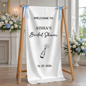 Könnte beinhalten: Ein weißes Stoffbanner auf einer Holzstaffelei, mit hellblauen Schleifen oben. Auf dem Banner steht "WELCOME TO AISHA'S Bridal Shower" mit einer Champagnerflaschen-Illustration und dem Datum 12.07.2026.