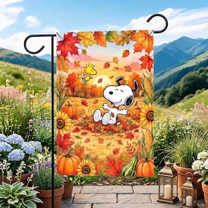 May include: A rectangular garden flag featuring a cartoon illustration of Snoopy sitting in a pile of autumn leaves with Woodstock flying above. The scene includes orange pumpkins, yellow sunflowers, corn stalks, and falling maple leaves in shades of red and gold.
