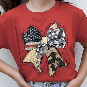 May include: Red t-shirt featuring a patriotic bow design. The bow incorporates the American flag, an eagle, and a soldier silhouette. The words "Honor the brave" and "Freedom Valor" are also visible.