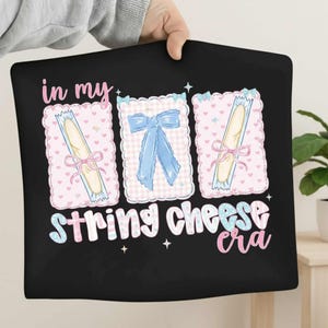 Könnte beinhalten: Schwarzes Kissen mit dem Text "in my string cheese era". Das Design zeigt drei illustrierte Käsepäckchen mit rosa Schleifen und einer blauen Schleife. Der Hintergrund ist rosa mit Herzen.