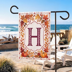May include: A rectangular garden flag featuring a central white scalloped frame with a large purple letter H. The border is decorated with a vibrant autumn pattern of orange and red maple leaves, acorns, berries, and two orange pumpkins.