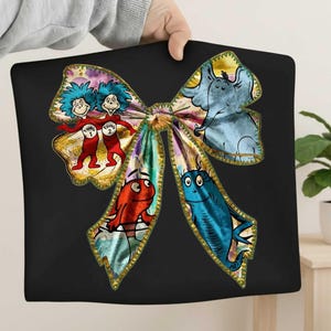 May include: Black pillow featuring a colorful bow design with illustrations of characters from a children's book. The bow includes images of Thing 1 and Thing 2, an elephant, a red fish, and a blue fish. The bow has a gold trim.