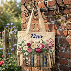 Peut inclure: Un sac fourre-tout matelassé de couleur crème orné d'une illustration imprimée d'une rangée de livres anciens surmontée d'un bouquet coloré de roses et de fleurs sauvages. Le prénom Eliza est inscrit en bleu au-dessus des fleurs.