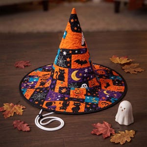 May include: A quilted witch hat featuring a vibrant Halloween-themed patchwork pattern with black cats, orange pumpkins, white spiderwebs, yellow crescent moons, and bats. The hat includes a black brim edge and a white adjustable chin strap.