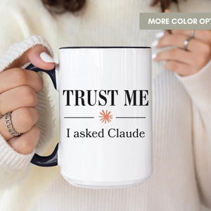 May include: A white ceramic mug with a dark blue handle and interior rim. The front features the black text "TRUST ME" above a small orange starburst icon, followed by the text "I asked Claude" in black. The mug has a cylindrical shape.