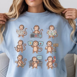 Puede incluir: Sudadera azul claro con un patrón repetido de monos de dibujos animados en varias poses. Los monos se representan con diferentes accesorios, como un periódico, una radio, un mapa y un teléfono. La sudadera está hecha de un material suave.