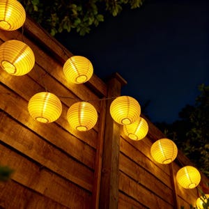 May include: A string of illuminated yellow paper lanterns hanging on a wooden fence at night. The round lanterns are evenly spaced and emit a warm glow, creating a festive atmosphere. The wooden fence is made of vertical planks.