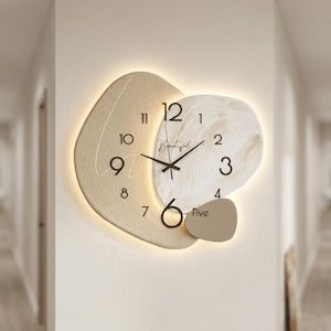 May include: A contemporary wall clock with an abstract design. The clock has a beige, irregular shape with a white and beige marbled section. The clock hands are black, and the numbers are in a simple font. The word "Beautiful" is written on the clock face.