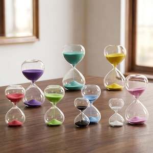 May include: A selection of clear glass hourglasses filled with coloured sand. The sand colours include red, purple, green, blue, yellow, pink, and black. The hourglasses are displayed on a wooden surface.