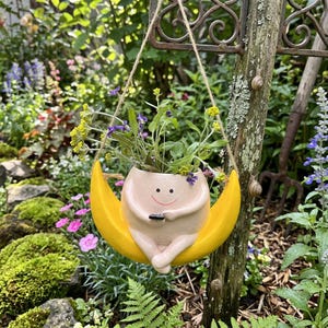 Puede incluir: Una maceta colgante caprichosa con forma de figura sonriente sentada en una luna creciente amarilla. La maceta está llena de vegetación y pequeñas flores moradas y amarillas. Se suspende con una cuerda natural.