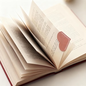 May include: An open book with pages fanned out, featuring a heart-shaped, pink leather bookmark with white stitching. The book has a red cover and the pages are filled with printed text.