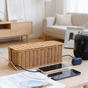 Puede incluir: Un organizador de cables rectangular de mimbre tejido de color marrón natural se encuentra sobre una mesa de madera. Dos teléfonos inteligentes se están cargando, conectados por cables. También se ve un estuche de carga azul y un altavoz negro.