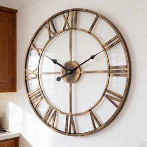 Puede incluir: Reloj de pared grande y redondo con marco de metal color bronce y números romanos. El reloj tiene un acabado envejecido, manecillas negras y una esfera central circular. El reloj está montado en una pared blanca.