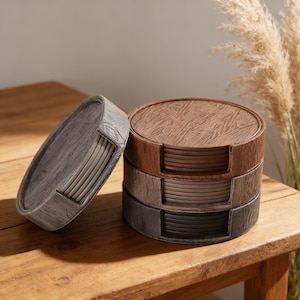 Puede incluir: Un juego de cuatro posavasos redondos en un soporte, con un diseño de vetas de madera. Los posavasos son en tonos de gris, marrón y negro. El soporte también es de vetas de madera, con una tapa a juego.