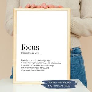 May include: A framed print featuring the word "focus" in a black, modern font, with its definition below. The frame is light brown. A blue banner at the bottom states "DIGITAL DOWNLOAD NO PHYSICAL ITEMS". The print is held by a person.