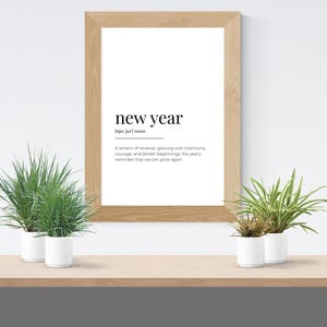 May include: A framed print with the words "new year" in a minimalist design. The definition of "new year" is below the title. Two small potted plants sit on a wooden shelf below the print.