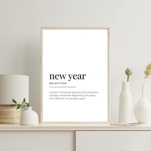 May include: A framed print with the words "new year" in a minimalist font. The definition below describes renewal and growth. The print is displayed on a white shelf with a lamp, vases, and decorative plants.