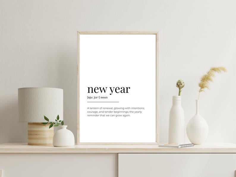 May include: A framed print with the words "new year" in a minimalist font. The definition below describes renewal and growth. The print is displayed on a white shelf with a lamp, vases, and decorative plants.