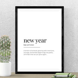 May include: A framed print with the words "new year" in a minimalist design. The text includes a definition and a motivational message. A small cactus in a white pot sits to the left of the frame. A stack of notebooks is at the bottom.