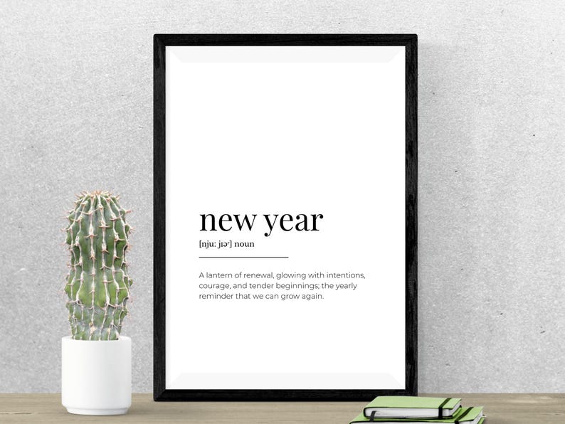 May include: A framed print with the words "new year" in a minimalist design. The text includes a definition and a motivational message. A small cactus in a white pot sits to the left of the frame. A stack of notebooks is at the bottom.