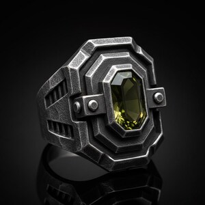 May include: A silver-toned ring with a geometric design and a rectangular, olive-green gemstone. The ring has a textured, industrial aesthetic, with a dark background. The gemstone is set within a detailed, layered bezel.