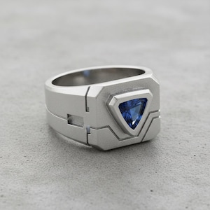 May include: A silver-colored ring with a geometric design. The ring features a triangular, deep blue gemstone set in a square face. The band has a unique, layered construction, giving it a modern, industrial aesthetic.