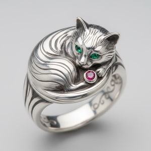 May include: A silver ring featuring a curled-up cat design. The cat has green gemstone eyes and a small pink gemstone held between its paws. The ring has a detailed, textured surface.