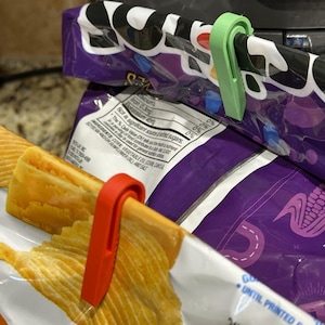 May include: Close-up of a bag of chips secured with a red and a green plastic clip. The chips are visible, with a yellow and orange color. The bag is purple and white, with nutritional information visible.