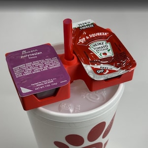 May include: A red plastic sauce holder with two compartments, holding a packet of Heinz tomato ketchup and a packet of Chick-fil-A Polynesian sauce. The holder sits atop a white cup with red polka dots.